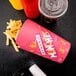 A pink Solo paper container of french fries and a drink on a table with a red bottle and a plastic cup with a lid.