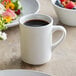 An Acopa ivory stoneware coffee mug filled with coffee on a table with a bowl of fruit.