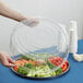 A person holding a clear plastic round high dome lid over a tray of vegetables.