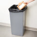 A person's hand putting a brown cardboard box into a gray Continental Swingline trash can.