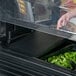 A hand opening a black Cambro Versa Well cover over a tray of greens on a salad bar.