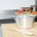 A silver Vollrath Arkadia sauce pan with a lid and a wooden spoon on a wooden table.