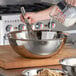 A hand pouring milk into a Vollrath stainless steel mixing bowl with a whisk.