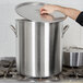 A person's hand opening a large Vollrath stainless steel stock pot on a stove.