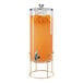 A Cal-Mil round glass beverage dispenser with an infusion chamber and brass wire base filled with orange juice.