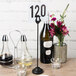 A black American Metalcraft harp card holder on a table with a number and flowers.