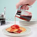 A hand using a Tablecraft Modern Glass Syrup Dispenser to pour syrup over pancakes.