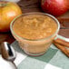 A glass bowl of Kime's sweetened cinnamon applesauce with a spoon and apples on a table.