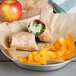 A plate with a burrito and chips next to an apple and a plate of food with a fruit and a plate of chips on a table.