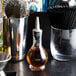 A clear glass American Metalcraft bitters bottle on a table next to a glass of brown liquid.
