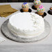 A white cake on a silver Enjay cake drum with cupcakes on a table.