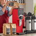A person in a red apron using a Bunn Bulk Coffee Grinder to fill a brown bag with coffee.