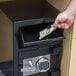 A person's hand putting a dollar bill into a FireKing black depository security safe.