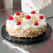 A cake with whipped cream and cherries on a black round cake board sitting on a table.