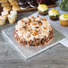 A cake with yellow frosting and nuts on a silver Enjay square cake drum on a table in a bakery display.