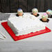 A white frosted cake on a red Enjay cake drum on a table with cupcakes and sprinkles.