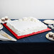 A white square cake with red frosting on a table with a red cake board underneath.