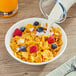 A bowl of Corn Flakes cereal with berries and milk on a table with a spoon and a napkin.