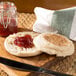 A buttered Thomas' Original English Muffin with jam on a wooden board with a knife and jar of jam.