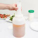 A hand pouring liquid from a plastic bottle with a white cap into a white Carlisle Store 'N Pour container.