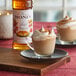 A glass cup of coffee with whipped cream and Monin syrup on a table with a bottle of Monin syrup.