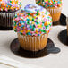 A close up of a cupcake with sprinkles on an Enjay black and gold reversible round dessert board with a tab.