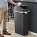 A man throwing a white cup into a black Lavex Slim Rectangular Trash Can with a swing dome lid.