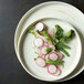 A Oneida Marble porcelain plate with a raised rim holding radishes and peas on a table with other vegetables and greens.