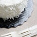 An Enjay black laminated corrugated cake circle under a white cake with white frosting on a black plate with plastic forks.