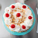 A blue Enjay round cake drum under a cake with white frosting and red cherries.