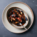 An oval bowl filled with mussels in sauce with a fork on the table.
