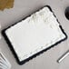 A white cake with frosting on a black Enjay 1/4 sheet cake board on a table.