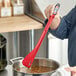 A person using a red Matfer Bourgeat Exoglass spatula to stir soup on a stove.