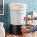 A round 6-gallon white beverage dispenser with a black base being used to pour a drink into a plastic cup.