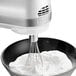 A KitchenAid white hand mixer with whisk attachment mixing whipped cream in a bowl.