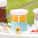 A table with a Choice translucent beverage dispenser with a blue base filled with a drink and a bowl of blackberries.