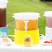 A Choice translucent plastic beverage dispenser with yellow base on a table with cups and a glass jar of pink liquid.