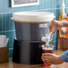 A round, translucent 3-gallon beverage dispenser with a black base and a white lid, labeled for coffee, being used to pour a drink into a plastic cup.