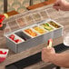 A person holding a stainless steel American Metalcraft condiment bar with fruit and drinks.