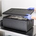 A person wearing blue gloves holding a black Cambro GoBox lid over an open container.
