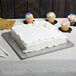 A white cake on a silver square cake drum with cupcakes on top.