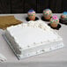 A white cake with frosting on a silver square cake drum with cupcakes on the table.