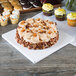 A white square cake drum with a frosted cake and cupcakes on a table.