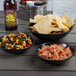 A black melamine serving bowl filled with corn and beans on a table with a bowl of chips and a bowl of food.