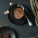 A Oneida Urban black porcelain saucer with a cup of hot chocolate and a spoon.