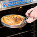 A hand using an American Metalcraft steel pizza pan gripper to hold a pizza in a pan.