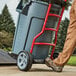 A man pushing a large grey Rubbermaid trash can on a dolly.