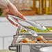 A person using American Metalcraft stainless steel serving tongs over a food tray.