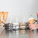 A hand holding a spoon over a waffle cone with a scoop of ice cream in a Tablecraft black powder coated condiment caddy.