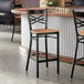 A black metal bar stool with a cross back design and a natural wood seat.